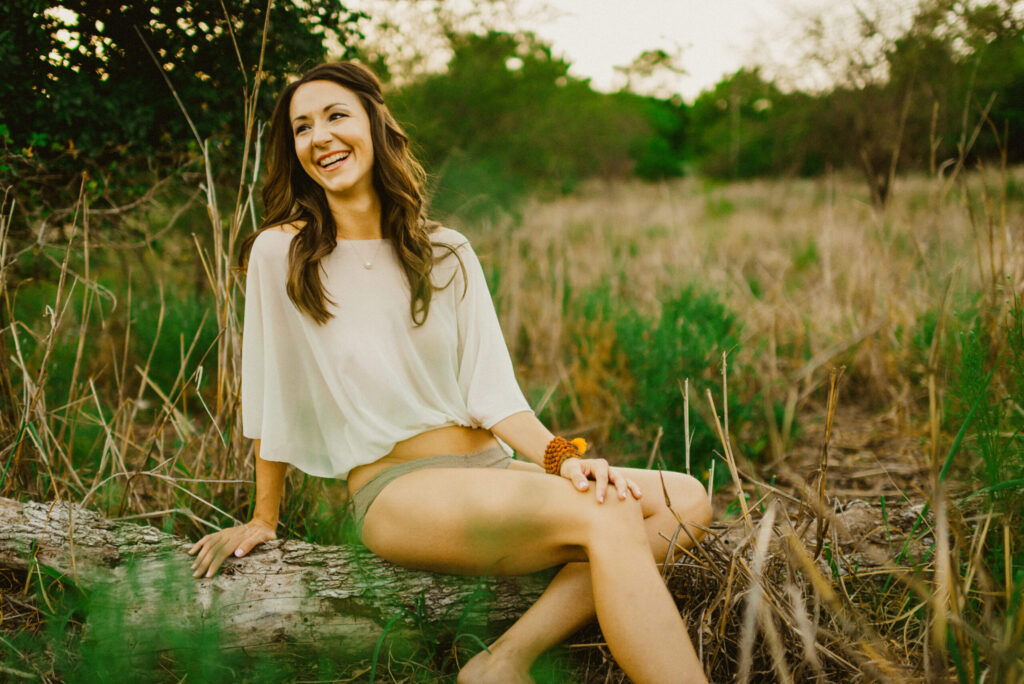 Woman smiling, sitting on log in grassy field.