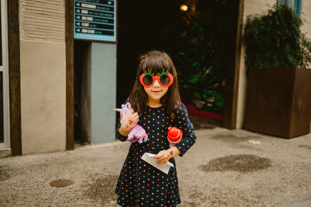 Child with sunglasses holding toy unicorn, polka dot dress.